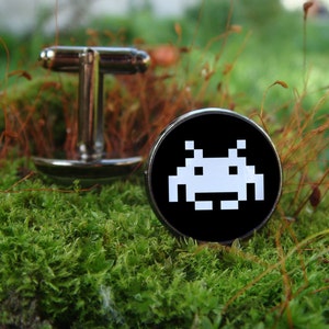 May include: A pair of silver cufflinks with a black background and a white pixelated alien design. The cufflinks are on a bed of green moss.