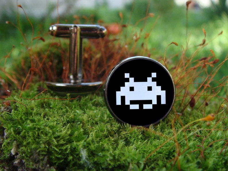 May include: A pair of silver cufflinks with a black background and a white pixelated alien design. The cufflinks are on a bed of green moss.