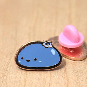 May include: A blue enamel pin shaped like a blob with a small white flower on top. The pin has a happy face and is attached to a gold-coloured backing.