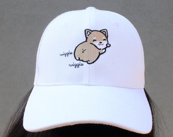 May include: White baseball cap featuring an embroidered cartoon Shiba Inu dog with the text "wiggle wiggle". The dog is light brown and cream-colored. The cap has a curved brim and a structured crown.