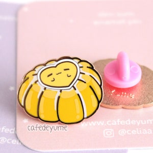 Könnte beinhalten: Eine goldfarbene Emaille-Anstecknadel mit einem gelben Cartoon-Pudding mit herzförmiger Oberseite und einem niedlichen Gesicht. Die Nadel hat auf der Rückseite einen rosa Plastik-Schmetterlingsverschluss.