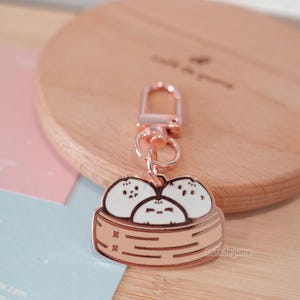 Könnte beinhalten: Rosenfarbener Schlüsselanhänger aus Metall mit drei Cartoon-Knödeln in einem Bambus-Dampfkorb. Der Schlüsselanhänger hat einen Karabinerhaken.