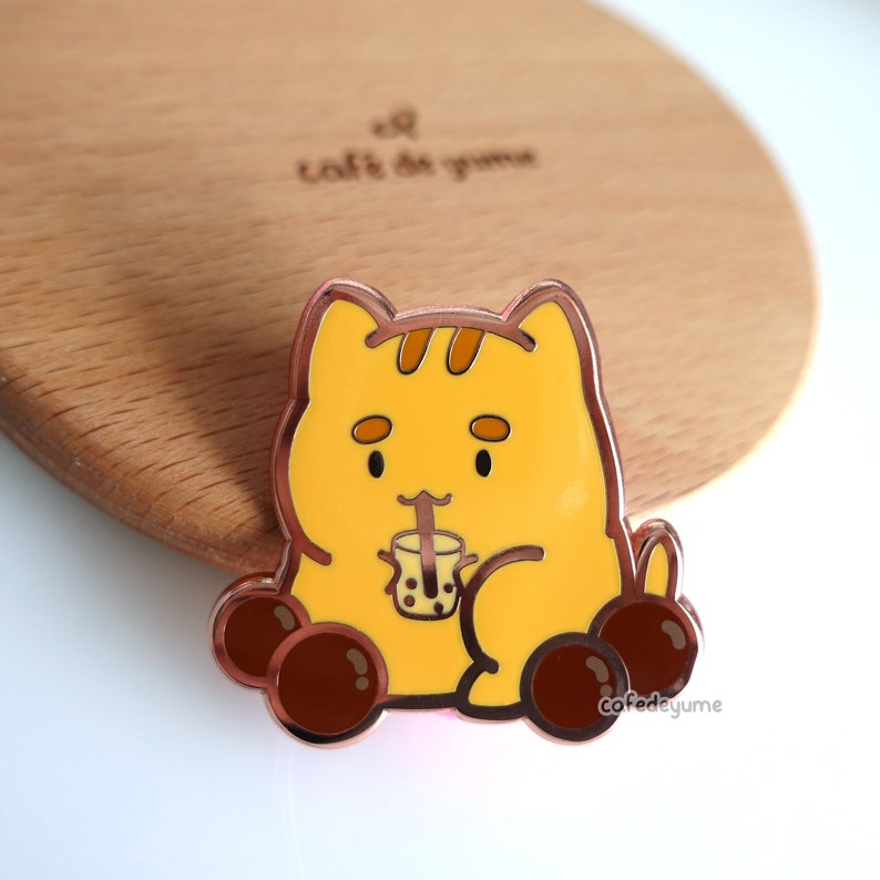 May include: A yellow cat-shaped enamel pin with brown accents, holding a bubble tea. The pin has a rose gold outline and brown bobbles. The text "cafedeyume" is on the bottom right. The pin is on a wooden surface.