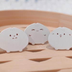 Könnte beinhalten: Drei weiße Keramikfiguren in Form von gedämpften Brötchen mit verschiedenen Gesichtsausdrücken. Die Brötchen stehen auf einer Holzoberfläche.