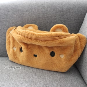 May include: A brown plush fanny pack shaped like a bear, featuring a zippered closure and embroidered details for eyes, nose, and heart-shaped cheeks. The bag has two small ears on top.