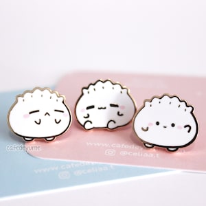 Könnte beinhalten: Drei goldfarbene Emaille-Pins in Form von weißen Knödeln mit niedlichen Comic-Gesichtern. Die Pins befinden sich auf einem rosa und blauen Hintergrund mit dem Text "www.cafedeyume.com @celiaa.t".
