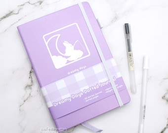 Może przedstawiać: Fioletowy notes z białym wzorem królika i księżyca. Notes ma biało-czarną wstążkę w kratkę i napis "dreamy days" oraz "Dreamy Days Dotted Journal" nadrukowany na okładce. Notes jest w komplecie z czarnym i białym długopisem.
