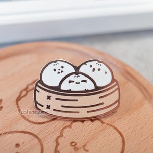 Könnte beinhalten: Eine goldfarbene Emaille-Anstecknadel mit drei Cartoon-Dampfbrötchen in einem Bambus-Dampfgarer. Die Brötchen haben süße Gesichter und sind weiß mit schwarzen Umrissen. Der Dampfgarer ist braun mit schwarzen Linien.