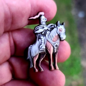 Op de afbeelding: Een zilveren en koperen emaille pin van een skeletridder die op een paard rijdt. De ridder draagt ​​een harnas en een helm met een pluim. Het paard heeft een witte manen en staart en een rood oog.
