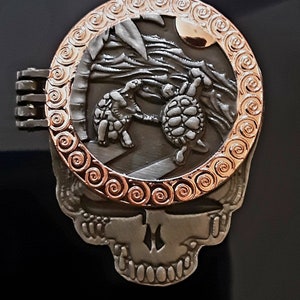 May include: A silver skull pendant with a round, rose gold locket featuring two turtles in a beach scene. The locket has a decorative, swirling border.