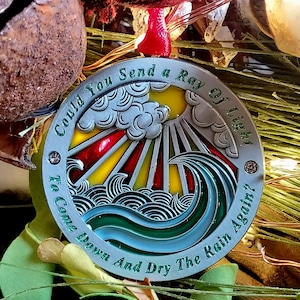 Puede incluir: Un adorno redondo de plata con un diseño de vitral de un sol brillando a través de nubes y olas. El adorno tiene el texto "Could You Send a Ray of Light To Come Down And Dry The Rain Again?" alrededor del borde.