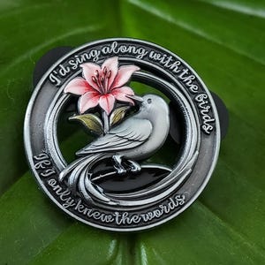 Op de afbeelding: Een ronde zilverkleurige metalen speld met een grijze vogel die op een tak naast een roze lelie zit. De buitenrand is gegraveerd met de tekst "I'd sing along with the birds" aan de bovenkant en "If I only knew the words" aan de onderkant.