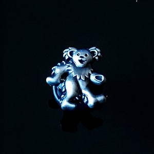 May include: A silver-colored bear figurine with a smiling face and a detailed design. The bear is standing with its arms raised, and it has a detailed collar. The figurine is set against a dark background.
