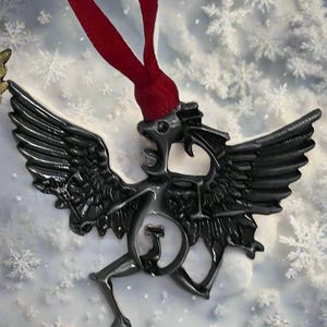 Puede incluir: Adorno de metal oscuro con forma de criatura alada con una cinta roja para colgar. El adorno tiene alas detalladas y un cuerpo estilizado. El fondo es blanco con adornos de copos de nieve.