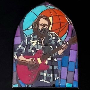 Può includere: Una finestra in vetro colorato che raffigura una persona che suona una chitarra elettrica rossa con un microfono di fronte a lei. Lo sfondo è un design in vetro colorato con tonalità blu, arancioni, viola e verdi.