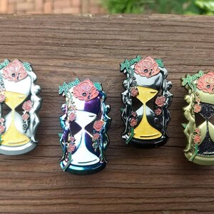 May include: Four enamel pins featuring an hourglass with a floral design. The hourglasses are in different colors: silver, purple, black, and gold. Each hourglass is surrounded by roses and vines.