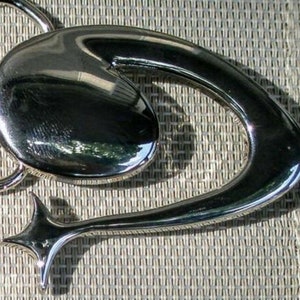 May include: A silver-toned metal belt buckle with a unique abstract design. The buckle features a large, rounded oval shape with a smaller, star-shaped design on the side.