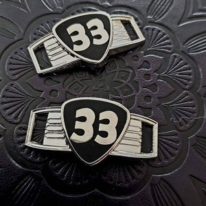 May include: Two metal emblems with a black shield shape and the number "33" in white. The emblems have a silver-colored frame with a rectangular design. The background is a dark, textured surface.