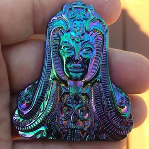 May include: A detailed, iridescent, metallic pin featuring a stylised, ornate, female figure with a crown and intricate details. The pin has a rainbow-like finish with shades of blue, green, purple, and gold.