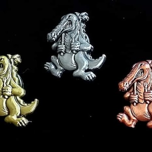 May include: Three metal alligator pins in gold, silver, and copper tones. Each pin depicts a cartoon alligator holding a fork and knife, with a running pose. The pins are set against a black background.