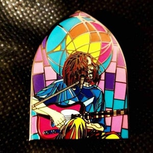 May include: A colorful enamel pin featuring a person playing a guitar in front of a stained glass window. The window is made of blue, purple, pink, and yellow glass.