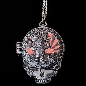 May include: A silver locket necklace with a detailed skull design. The locket opens to reveal a colorful image of a figure playing a musical instrument in front of a tree and a sunrise.