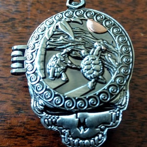 Puede incluir: Colgante de metal plateado en forma de calavera con un compartimento con bisagras. El colgante presenta una escena detallada de dos tortugas en una playa con palmeras y un sol color cobre. El diseño está rodeado por un borde decorativo en espiral.