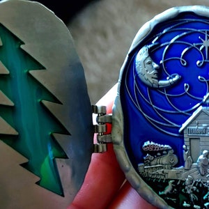 May include: A silver-toned, hinged, circular pendant with a green, stylized pine tree design on one side and a blue, swirling, celestial scene with a crescent moon, a star, and a small building on the other side. The building has the text "Terrapin Station" on the front.