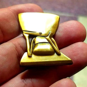May include: A gold-coloured metal pin shaped like a person sitting in a chair. The pin is detailed and has a smooth, shiny finish.