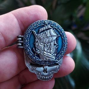 May include: A silver and blue metal pin with a detailed image of a ship sailing on a blue sea. The pin is shaped like a skull with a circular design around the ship.