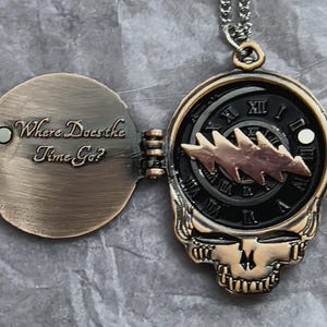 Könnte beinhalten: Eine bronzefarbene Anhängerkette mit Totenkopf-Design und einem Zifferblatt. Das Zifferblatt hat römische Ziffern und einen Blitz. Der Anhänger öffnet sich und zeigt den Text "Where Does the Time Go?"