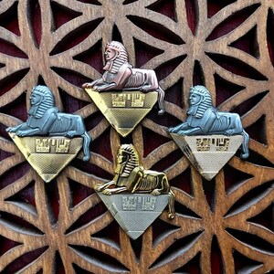 May include: Four decorative metal sphinx figurines with different colored finishes. Each sphinx is mounted on a triangular base with a gold, silver, or copper finish. The bases have the word "Egypt" printed on them.