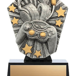May include: A silver-colored gaming trophy featuring a headset, a game controller, and gold stars. The trophy sits on a black base with a gold-colored plate for engraving. The design has a retro, arcade-like aesthetic.