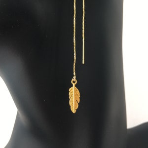 May include: Gold feather dangle earrings with a delicate chain. The earrings are on a black mannequin head.