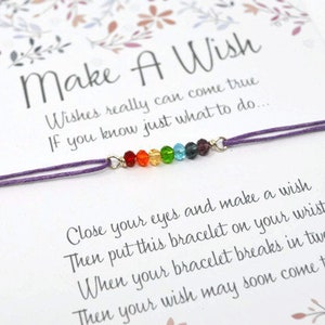 May include: A purple string bracelet with a row of rainbow-coloured beads, displayed on a card. The card reads "Make A Wish" and includes instructions for making a wish, with floral decorations.