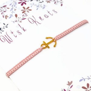 以下が含まれることがあります： ゴールドのアンカーチャームが付いたピンクの友情ブレスレット。ブレスレットは、「Wish Knots」というテキストが書かれた白いカードの上に置かれています。