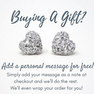 May include: Two silver heart-shaped stud earrings with a glittery texture. The image includes the text "Buying a Gift?" and "Add a personal message for free!"