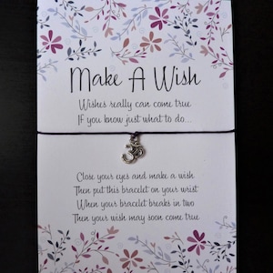 May include: A 'Make A Wish' card with a purple string bracelet featuring a silver charm. The card has floral designs in purple and pink. Text on the card reads: 'Wishes really can come true. If you know just what to do...'