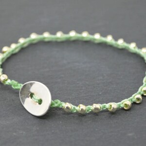 May include: A green and silver bracelet with a silver button closure. The bracelet is made of a thin cord with small silver beads.