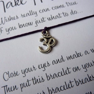 May include: A wish bracelet with a silver Om charm and a dark purple string. The bracelet is on a white card with text that reads "Make it Happen" and instructions for making a wish.