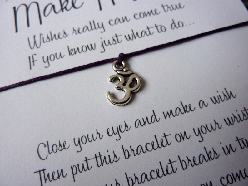 May include: A wish bracelet with a silver Om charm and a dark purple string. The bracelet is on a white card with text that reads "Make it Happen" and instructions for making a wish.
