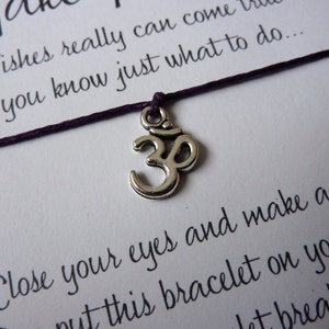 May include: A close-up of a wish bracelet with a purple cord and a silver Om charm. The card behind the bracelet has handwritten text that reads "Wishes really can come true if you know just what to do..." and "Close your eyes and make a wish."