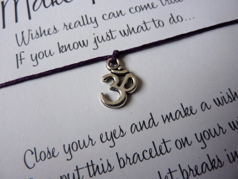 May include: A close-up of a wish bracelet with a purple cord and a silver Om charm. The card behind the bracelet has handwritten text that reads "Wishes really can come true if you know just what to do..." and "Close your eyes and make a wish."