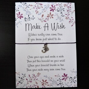 May include: A white card with the text "Make A Wish" and a purple string bracelet with a silver Om charm. The card has floral designs in purple and pink. The text on the card reads "Wishes really can come true..."
