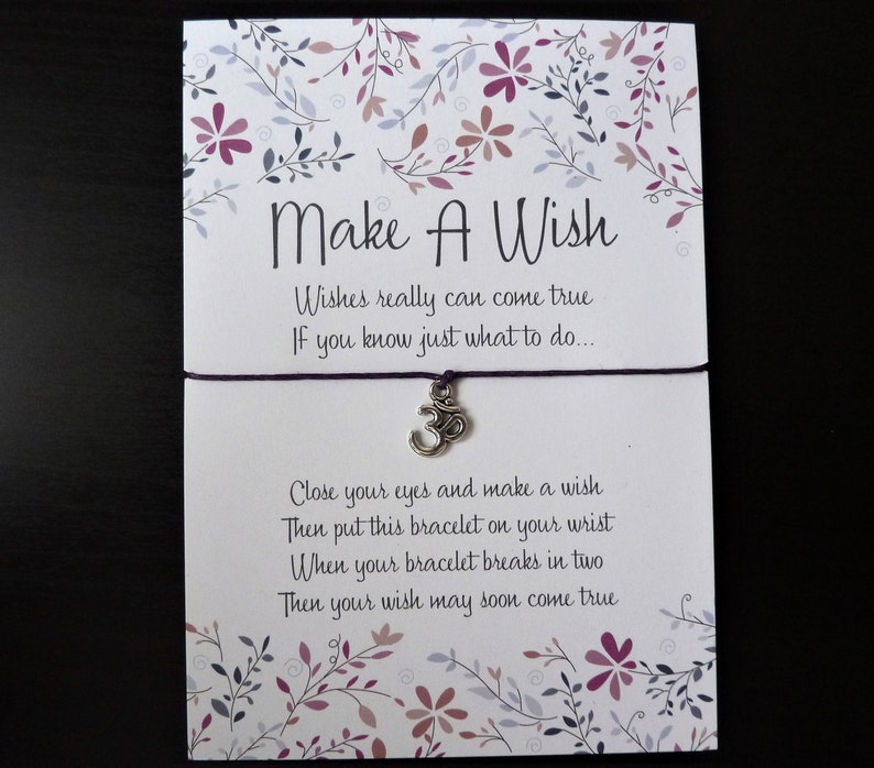 May include: A white card with the text "Make A Wish" and a purple string bracelet with a silver Om charm. The card has floral designs in purple and pink. The text on the card reads "Wishes really can come true..."
