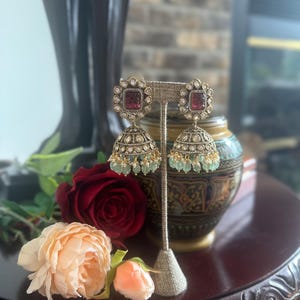 May include: A pair of ornate gold-toned earrings with a square, red gemstone center, surrounded by smaller clear stones. The earrings have a bell-shaped design with dangling green beads. The earrings are displayed on a stand with a dark wood table and flowers in the background.