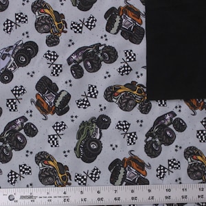 Peut inclure: Un tissu gris clair avec un motif répétitif de monster trucks colorés et de drapeaux à damier noir et blanc, agrémenté de petites étoiles noires. Une règle métallique de 30 cm est placée le long du bord inférieur.