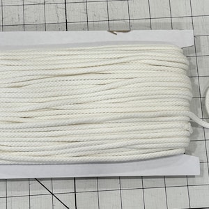 Peut inclure: Une bobine de corde blanche, bien enroulée et maintenue dans un support en carton. Une boucle de la corde s'étend de la bobine, suggérant son utilisation. La corde est d'un blanc vif et propre.