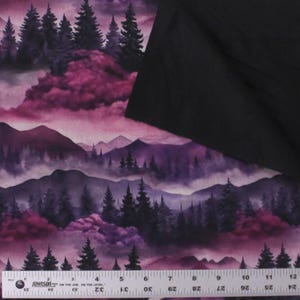 May include: A fabric featuring a printed landscape of dark pine trees and purple mountains under a pink and violet sky, partially covered by a solid black fabric swatch, with a ruler showing 12 inches at the bottom edge of the patterned material.