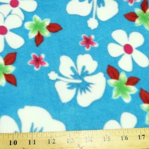 May include: A bright blue fleece fabric featuring a repeating pattern of large white hibiscus flowers with pink centers, smaller green and white star-shaped flowers, and dark red leaves. A wooden ruler is visible at the bottom edge.